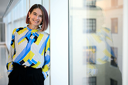 Zu sehen ist Prof. Dr. Catherine D'Ignazio in bunten Kleidern wie sie in einem hellen Raum an einem Balken neben Fenstern angelehnt ist.